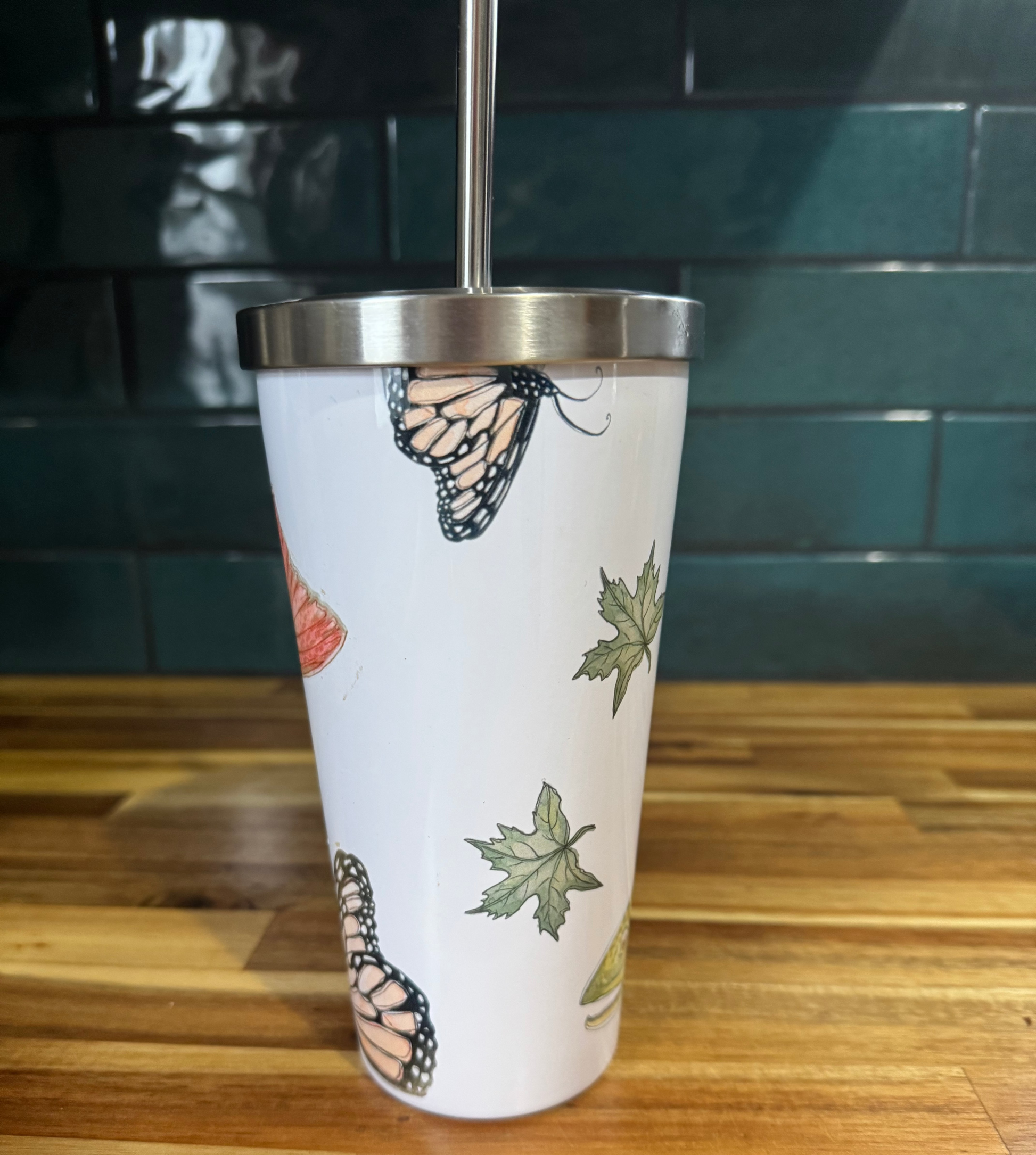 Tumbler with butterfly and leaf design on a wooden surface with a teal tiled wall in the background