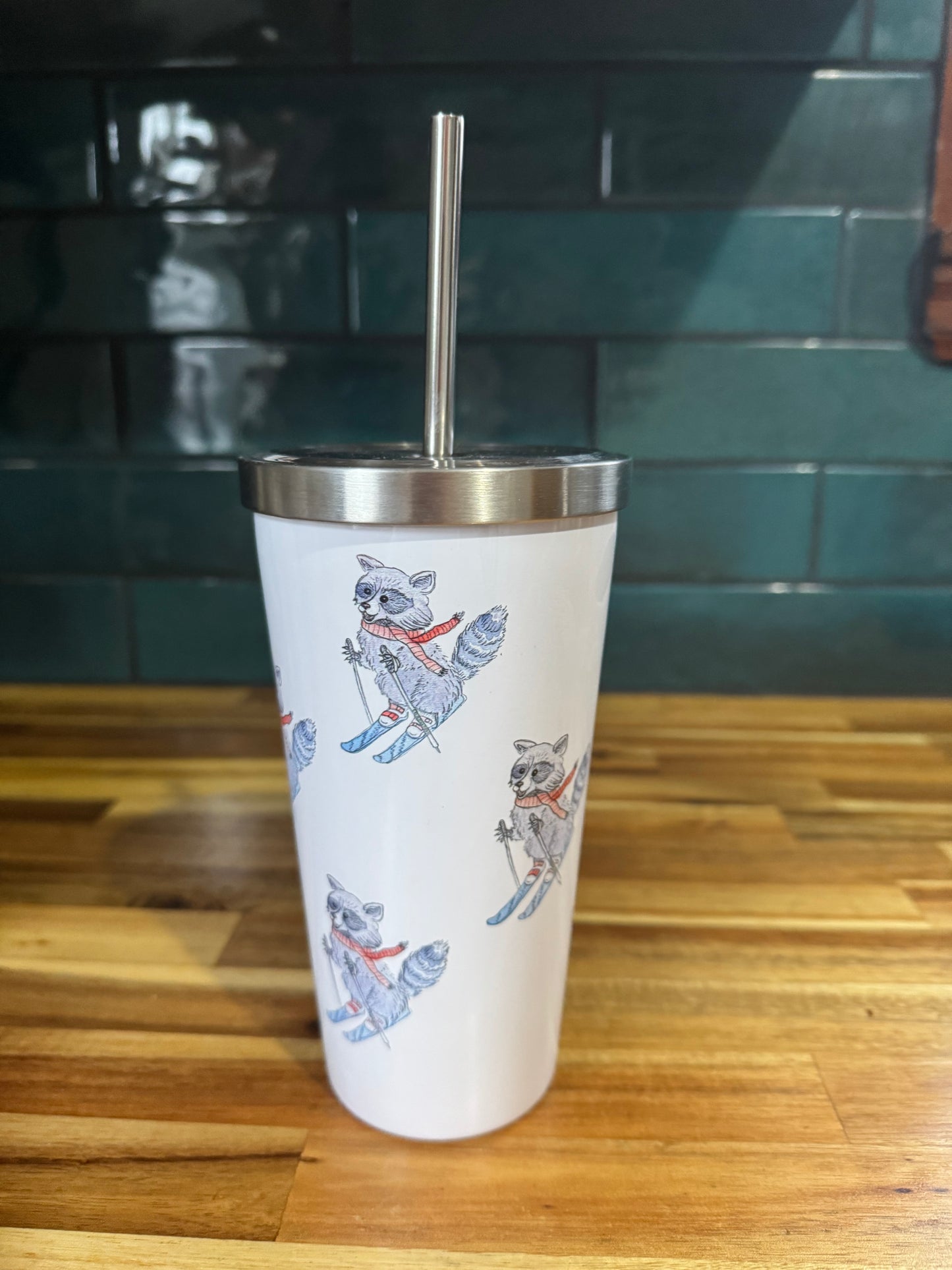 Tumbler with raccoon illustrations on a wooden surface and green tiled wall background