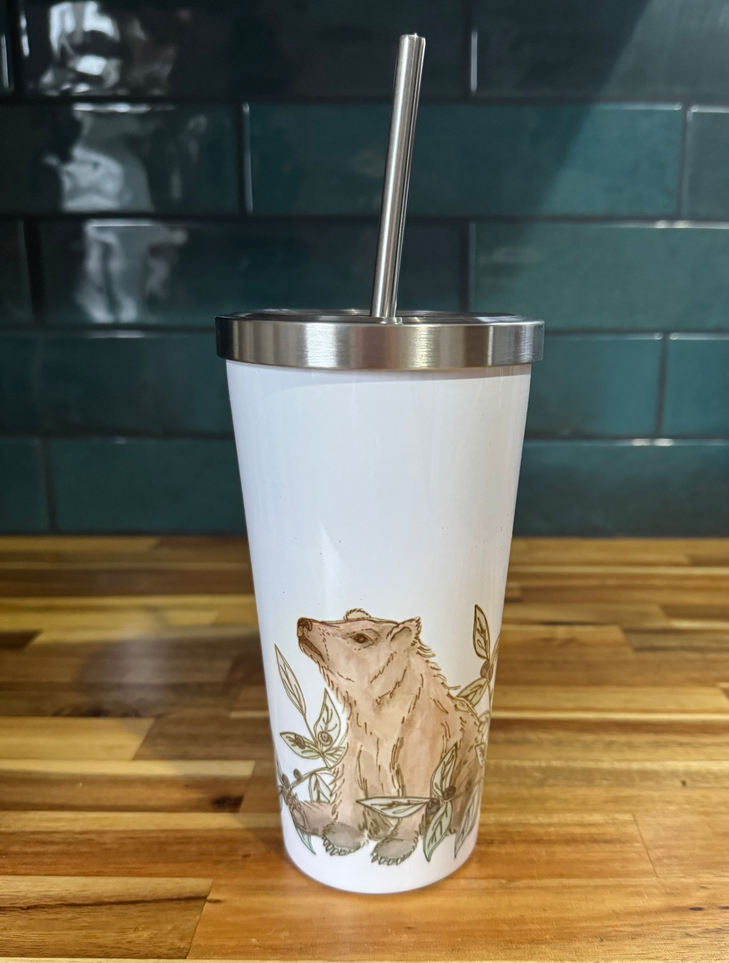 White tumbler with bear design and metal straw on a wooden surface